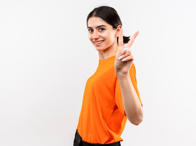Young girl wearing orange t-shirt smiling cheerfully showing v-sign standing over white wall