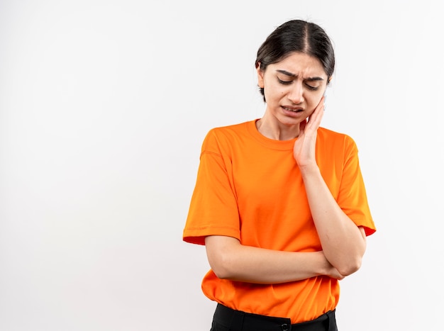 Young girl wearing orange t-shirt touching her cheek feeling toothache standing over white wall