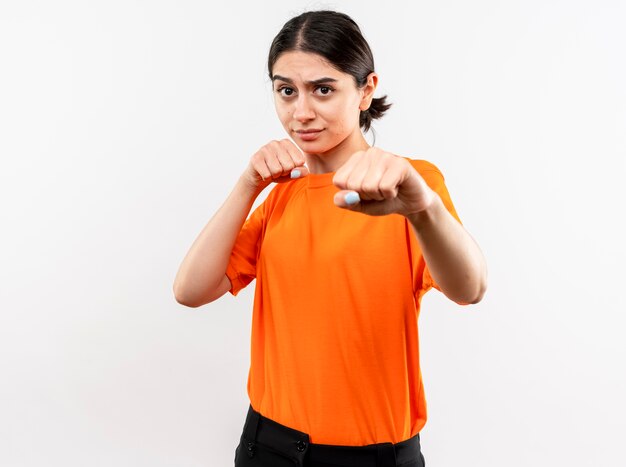 Young girl wearing orange t-shirt with clenched fist posing like a boxer standing over white wall