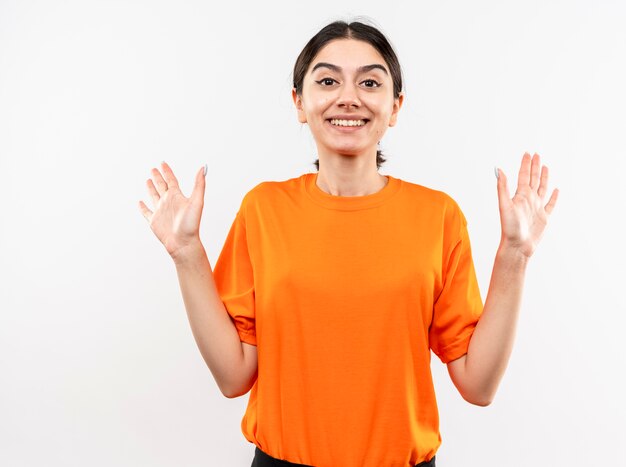 Молодая девушка в оранжевой футболке с поднятыми руками, весело улыбаясь, стоя над белой стеной