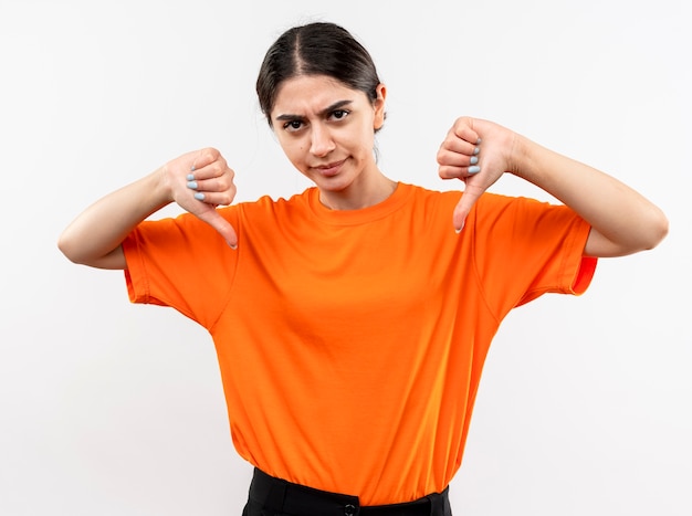 Free photo young girl wearing orange t-shirt with serious face frowning showing thumbs down standing over white wall