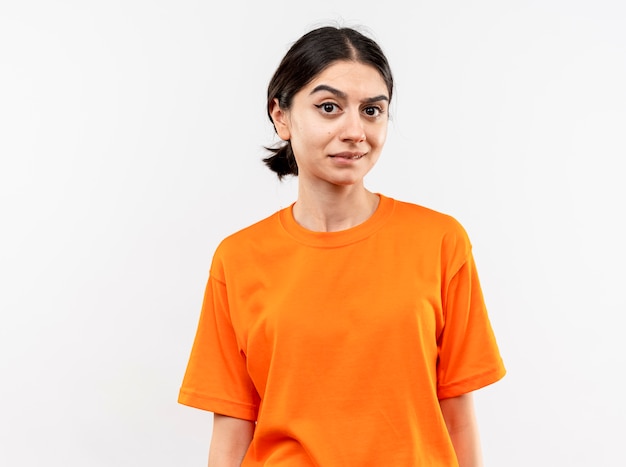 흰 벽 위에 서있는 회의적인 표정으로 오렌지 티셔츠를 입고 어린 소녀