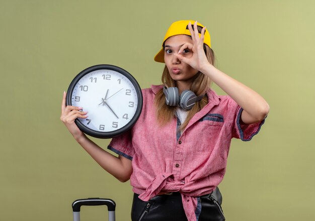 Young girl wearing pink shirt in cap with headphones around neck holding wall clock doing ik sing looking through this sign