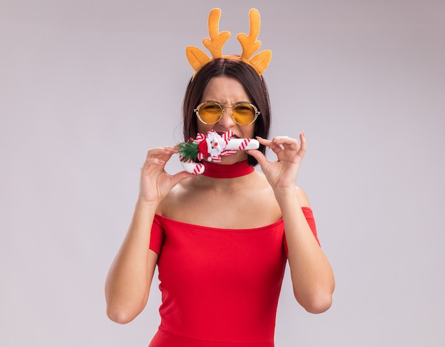 Young girl wearing reindeer antlers headband and glasses holding christmas candy cane ornament near mouth biting it looking at camera isolated on white background