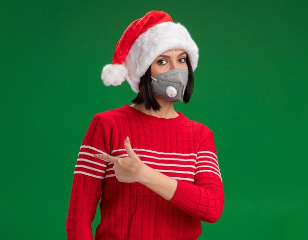 Young girl wearing santa hat and protective mask looking at camera pointing at side isolated on green background with copy space