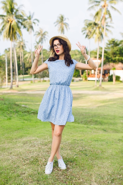 Young girl wearing sky-blue dress is standing on tip-toes in a park. Girl has straw hat and black glasses on