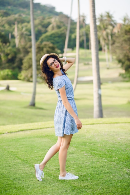 Young girl wearing sky-blue dress is standing in a tropical park. Girl has straw hat and black glasses on.