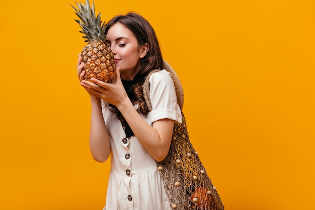 Young girl in white dress with shopping bag on her shoulder sniffing pineapple on orange background.