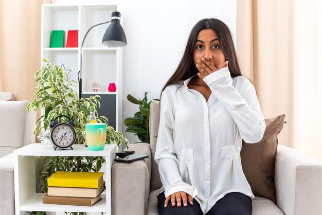 Young girl in white shirt and black pants  being in shocked covering mouth with hand sitting on the chair in light living room