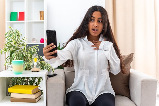 Young girl in white shirt and black pants holding smartphone looking at it confused and surprised sitting on the chair in light living room