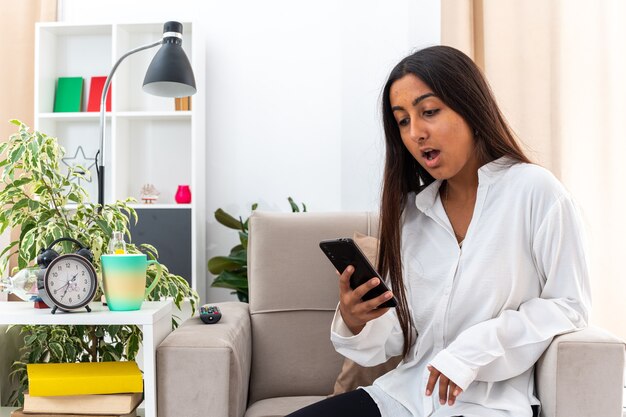 Young girl in white shirt and black pants holding smartphone looking at it surprised and very anxious sitting on the chair in light living room