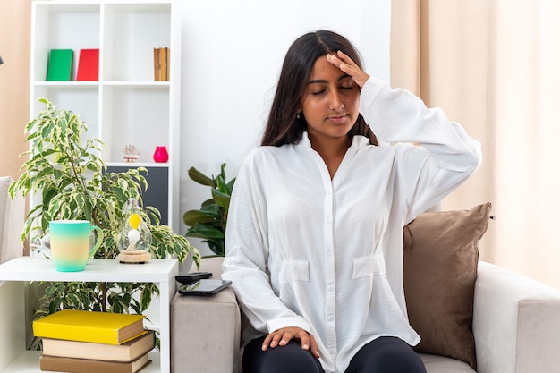 Young girl in white shirt and black pants looking unwell touching her head suffering from headache sitting on the chair in light living room