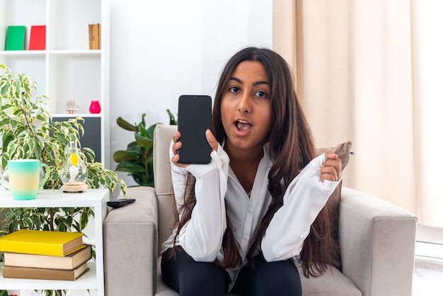 Young girl in white shirt and black pants showing smartphone looking happy and excited sitting on the chair in light living room