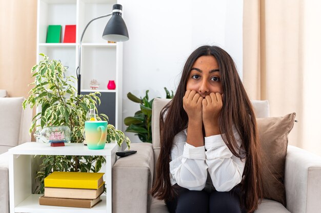 Young girl in white shirt and black pants  stressed and nervous biting nails sitting on the chair in light living room