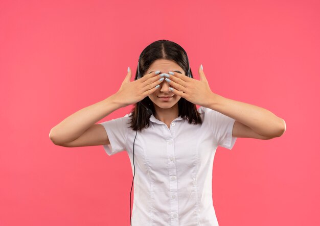 Young girl in white shirt and headphones, covering eyes with hands standing over pink wall