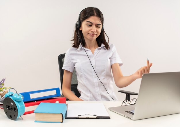 Young girl in white shirt and headphones, looking displeased sitting at the table with folders and laptop over white wall