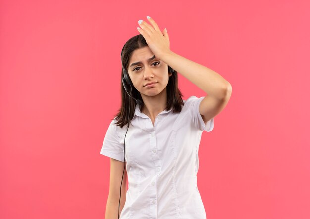 ピンクの壁の上に立っている間違いのために彼女の頭の上の手と混同された正面を見て、白いシャツとヘッドフォンの少女