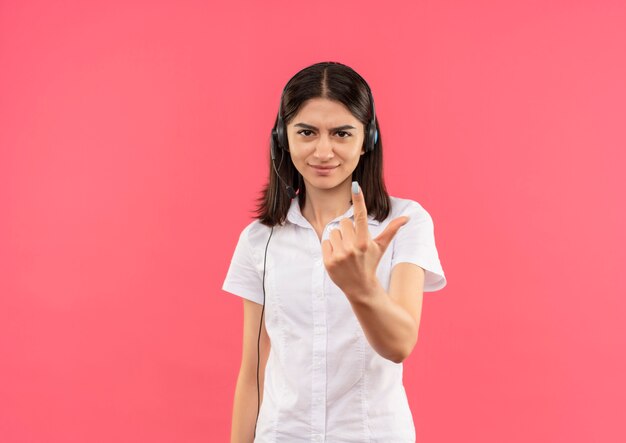 Young girl in white shirt and headphones, looking to the front displeased showing index finger standing over pink wall