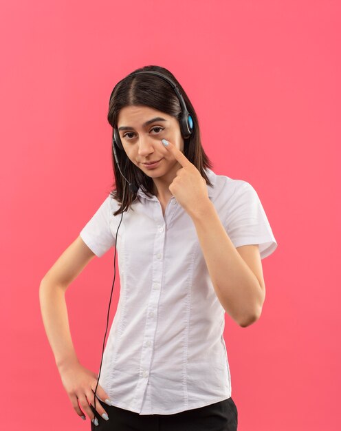 Young girl in white shirt and headphones, looking to the front pointing to her eye watching you gesture standing over pink wall