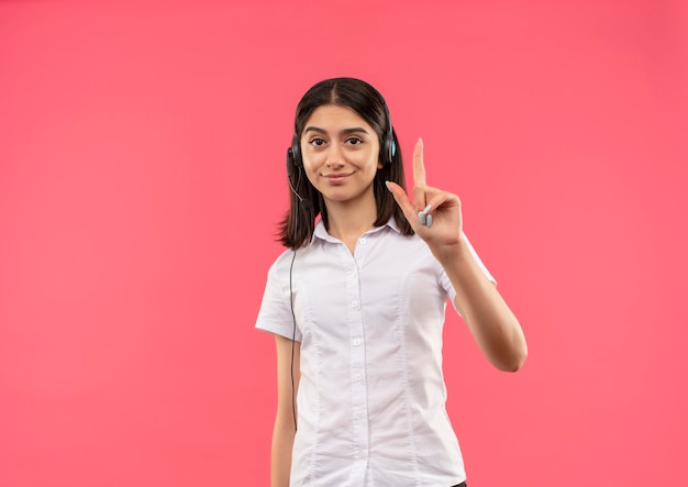 Young girl in white shirt and headphones, looking to the front smiling confident showing index finger standing over pink wall