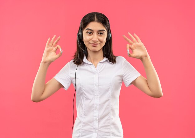 흰 셔츠와 헤드폰에 어린 소녀, 분홍색 벽 위에 서있는 양손으로 확인 표시를 보여주는 자신감 웃고 전면을 찾고