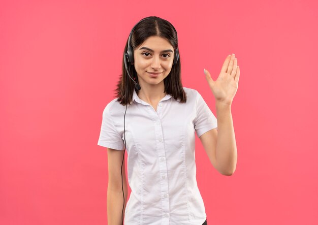 Young girl in white shirt and headphones, looking to the front smiling friendly waving with hand standing over pink wall