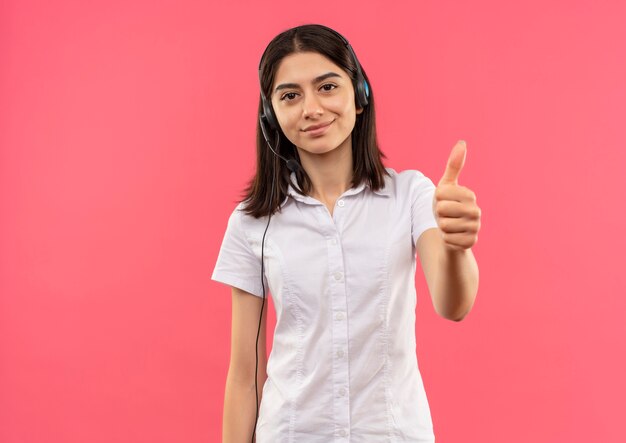 Young girl in white shirt and headphones, looking to the front smiling showing thumbs up standing over pink wall