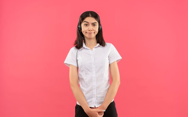 Young girl in white shirt and headphones, looking to the front with skeptic smile standing over pink wall