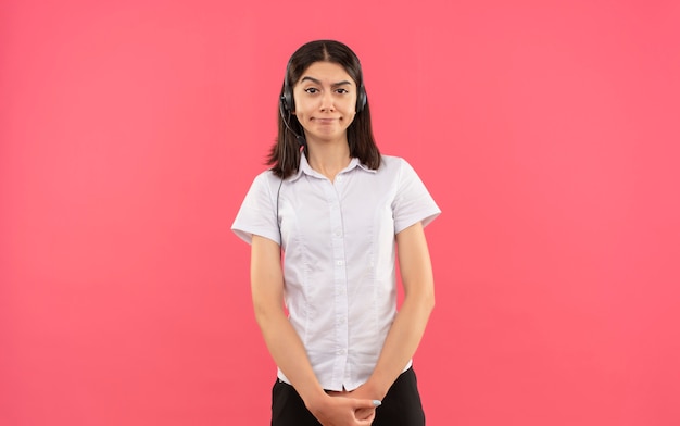 Молодая девушка в белой рубашке и наушниках, смотрящая вперед со скептической улыбкой, стоит над розовой стеной