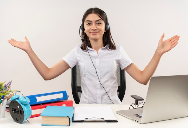 Foto gratuita giovane ragazza in camicia bianca e cuffie, guardando in avanti con il sorriso sul viso, diffondendo le mani ai lati seduti al tavolo con cartelle e laptop sul muro bianco