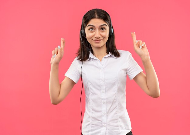 Young girl in white shirt and headphones, making desirable wish crossing fingers happy and positive standing over pink wall