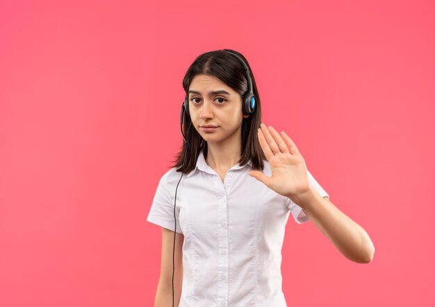 Young girl in white shirt and headphones, making stop gesture with hand as telling not to come closer standing over pink wall