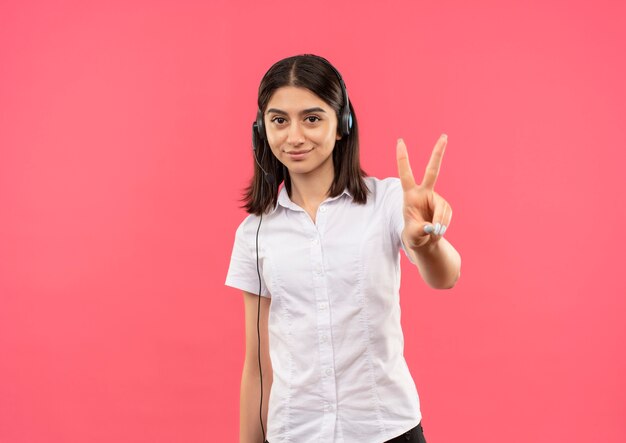 Молодая девушка в белой рубашке и наушниках, показывая пальцами номер два, улыбаясь стоя над розовой стеной