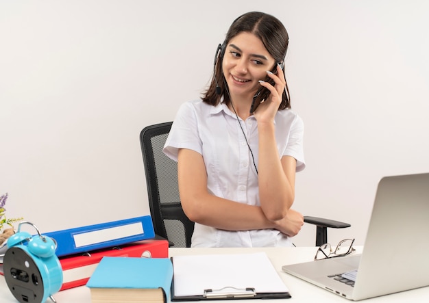 Foto gratuita giovane ragazza in camicia bianca e cuffie, parlando al telefono cellulare sorridente seduto al tavolo con cartelle e laptop sul muro bianco