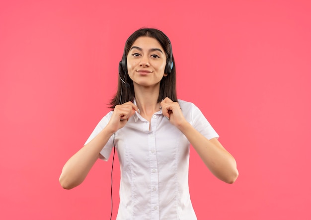 Foto gratuita giovane ragazza in camicia bianca e cuffie, toccando il suo collare cercando fiducioso in piedi sopra il muro rosa