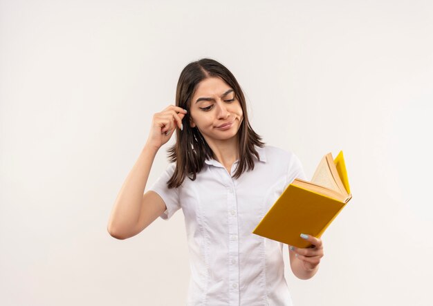 Young girl in white shirt holding book looking at it confused standing over white wall