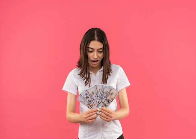 Young girl in white shirt holding cash looking amazed and surprised standing over pink wall