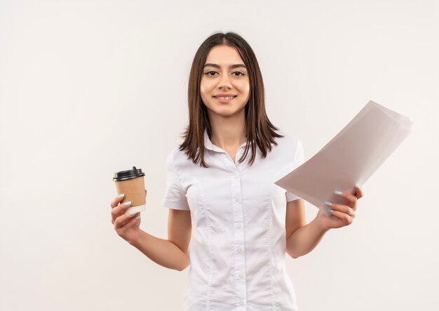 Young girl in white shirt holding coffee cup and blank pages smiling confident standing over white wall