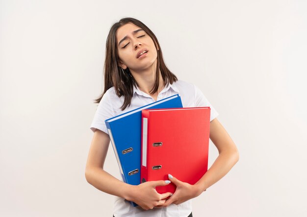 Young girl in white shirt holding folders looking tired and bored standing over white wall