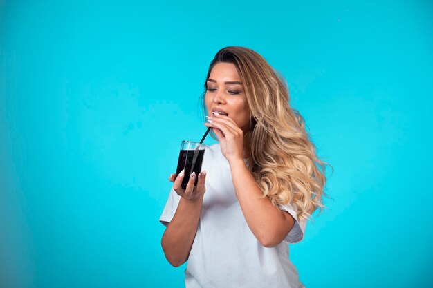 Молодая девушка в белой рубашке держит стакан черного коктейля и проверяет вкус на трубке.