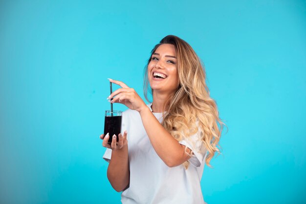 Молодая девушка в белой рубашке держит стакан черного коктейля и проверяет вкус.