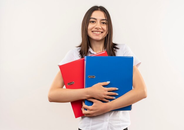 Young girl in white shirt holding two folders looking to the front smiling cheerfully standing over white wall