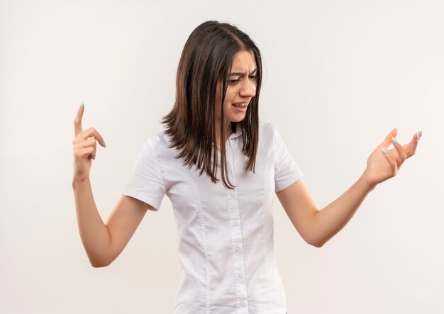 Young girl in white shirt looking aside disappointed with raised arms standing over white wall