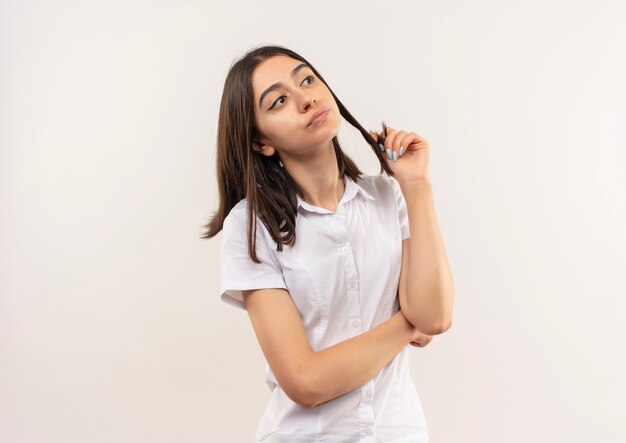 Молодая девушка в белой рубашке, озадаченная глядя в сторону, стоит над белой стеной