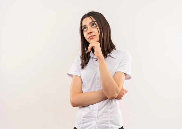 Young girl in white shirt looking aside with hand on chin puzzled standing over white wall