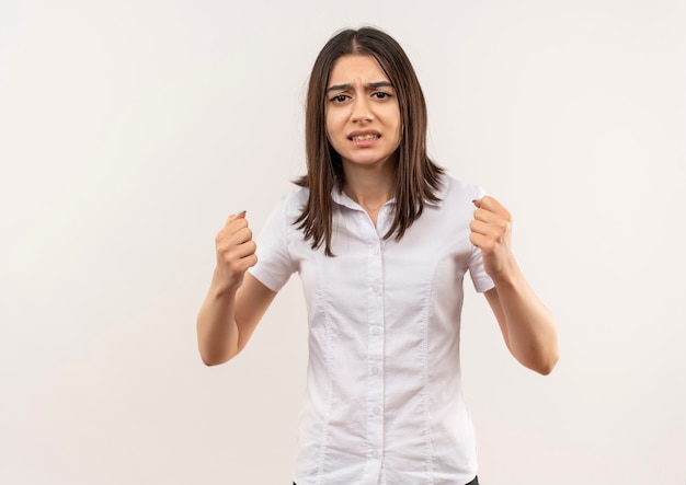 Young girl in white shirt looking to the front clenching fist with angry face standing over white wall