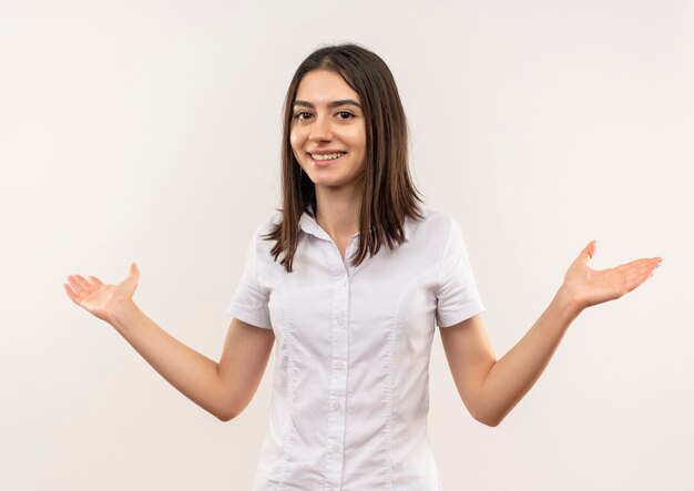 Young girl in white shirt looking to the front smiling cheerfully spreading arms to the sides standing over white wall