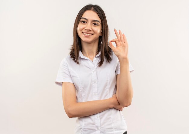 白い壁の上に立っているOKのサインを示して笑顔で正面を向いている白いシャツの少女