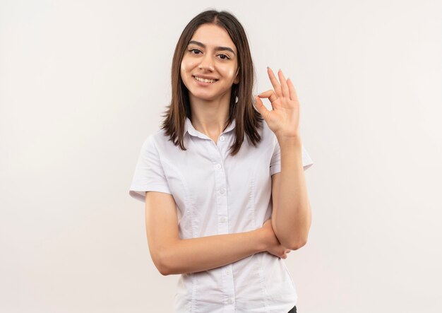 Young girl in white shirt looking to the front smiling showing ok sign standing over white wall