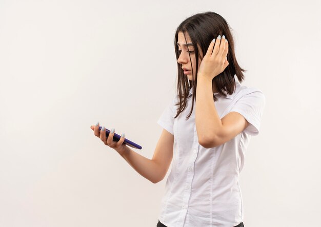 Young girl in white shirt looking at screen of her mobile phone confused standing over white wall